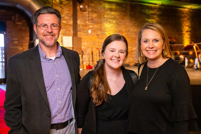 A man wearing a black blazer and two women wearing black outfits smile